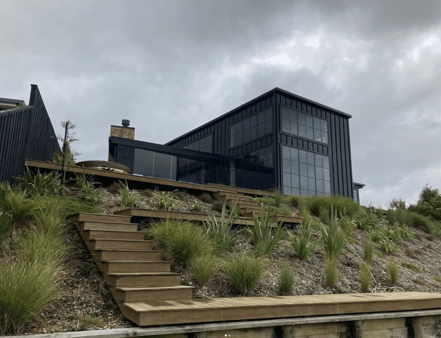 Modern black building with large glass windows, seen from a garden with wooden stairs and green plants